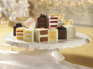 Variety of petits fours displayed on a white cake stand, cut to show the different layers.
