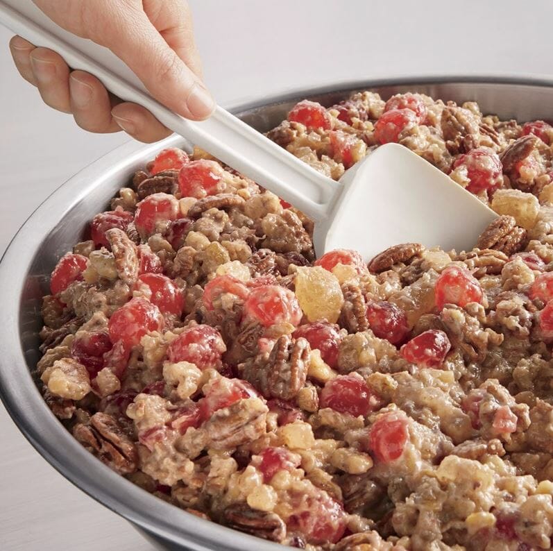 A hand holding a spatula stirring fruitcake nuts and candied fruits in a stainless bowl. 
