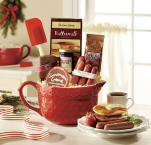A red mixing pitcher packed with pancake mix, syrup, breakfast links, coffee, apple bread, crème and a red spatula.