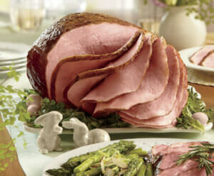 Sliced glazed Easter ham on a white platter, ceramic bunnies, and a side plate of asparagus and sliced  ham.