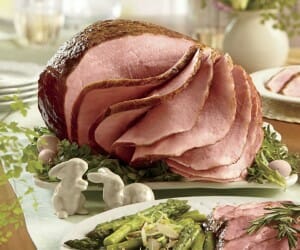 Sliced glazed Easter ham on a white platter, ceramic bunnies, and a side plate of asparagus and sliced  ham.