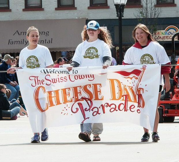 Three females at Cheese Days 2016 walking with a Swiss Colony Cheese Days parade banner.