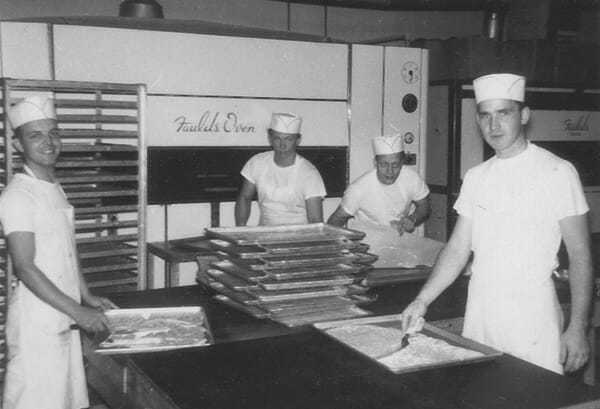 Four pastry chefs in white aprons and hats layering dobosh torte on pans in a pre-1970's bakery.
