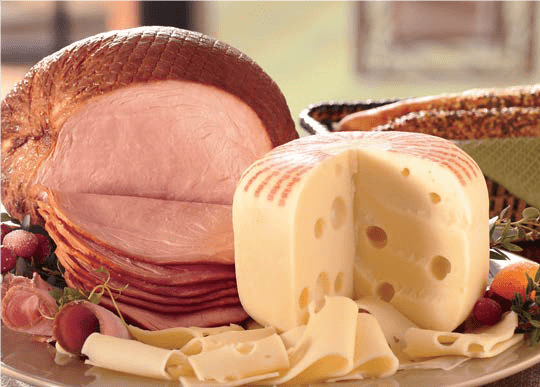 A glazed Sliced Ham next to a round of Baby Swiss Cheese, cut to show the holes, with slices on a platter.